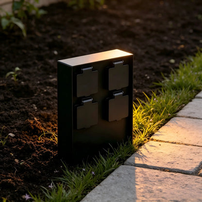 Black weatherproof 4-way outdoor power socket box installed in a garden flower bed along a stone path during sunset